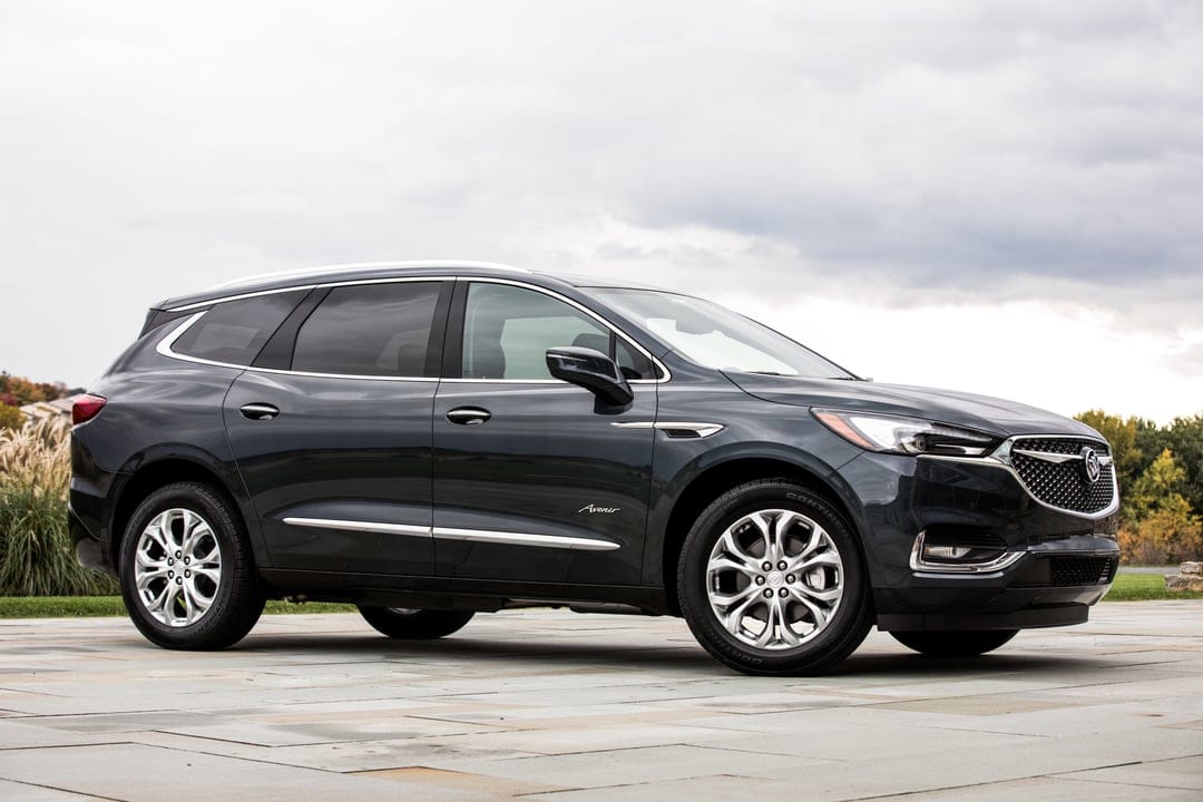 Side view of a dark gray Buick Enclave Avenir SUV on light stone pavers under an overcast sky with distant trees.