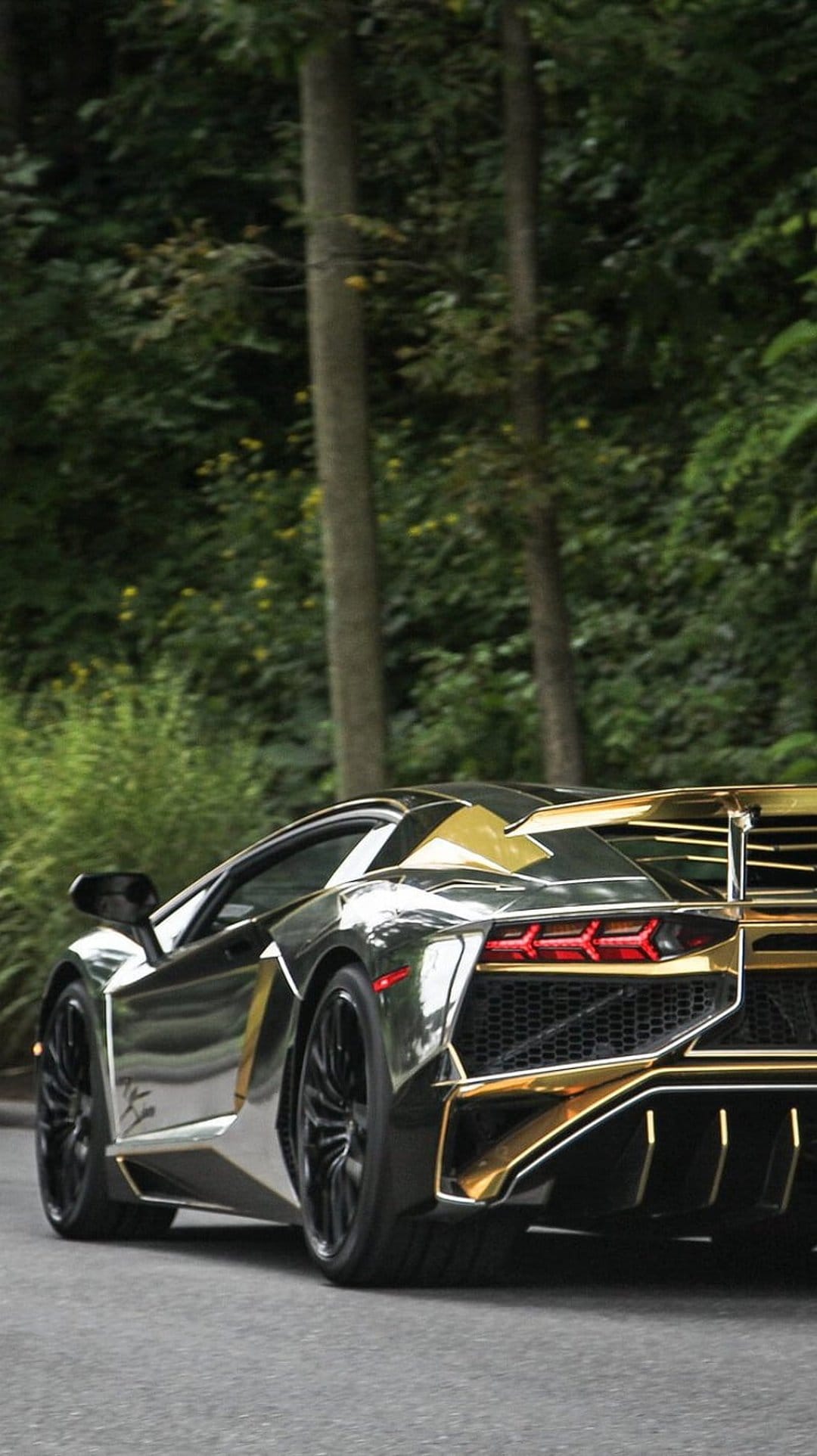 Gold-accented chrome Lamborghini Aventador rear view on a forest road surrounded by lush green trees and foliage.