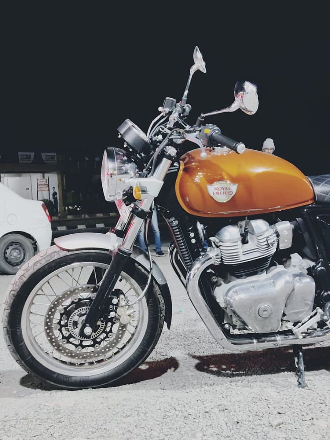 Royal Enfield motorcycle with an orange tank and chrome engine components parked in a dark nocturnal setting.
