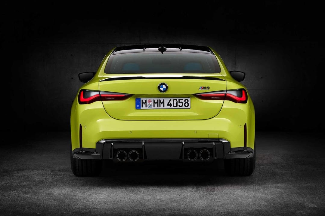 Rear view of a yellow BMW M4 Competition with glowing red taillights and quad exhausts against a dark wall.