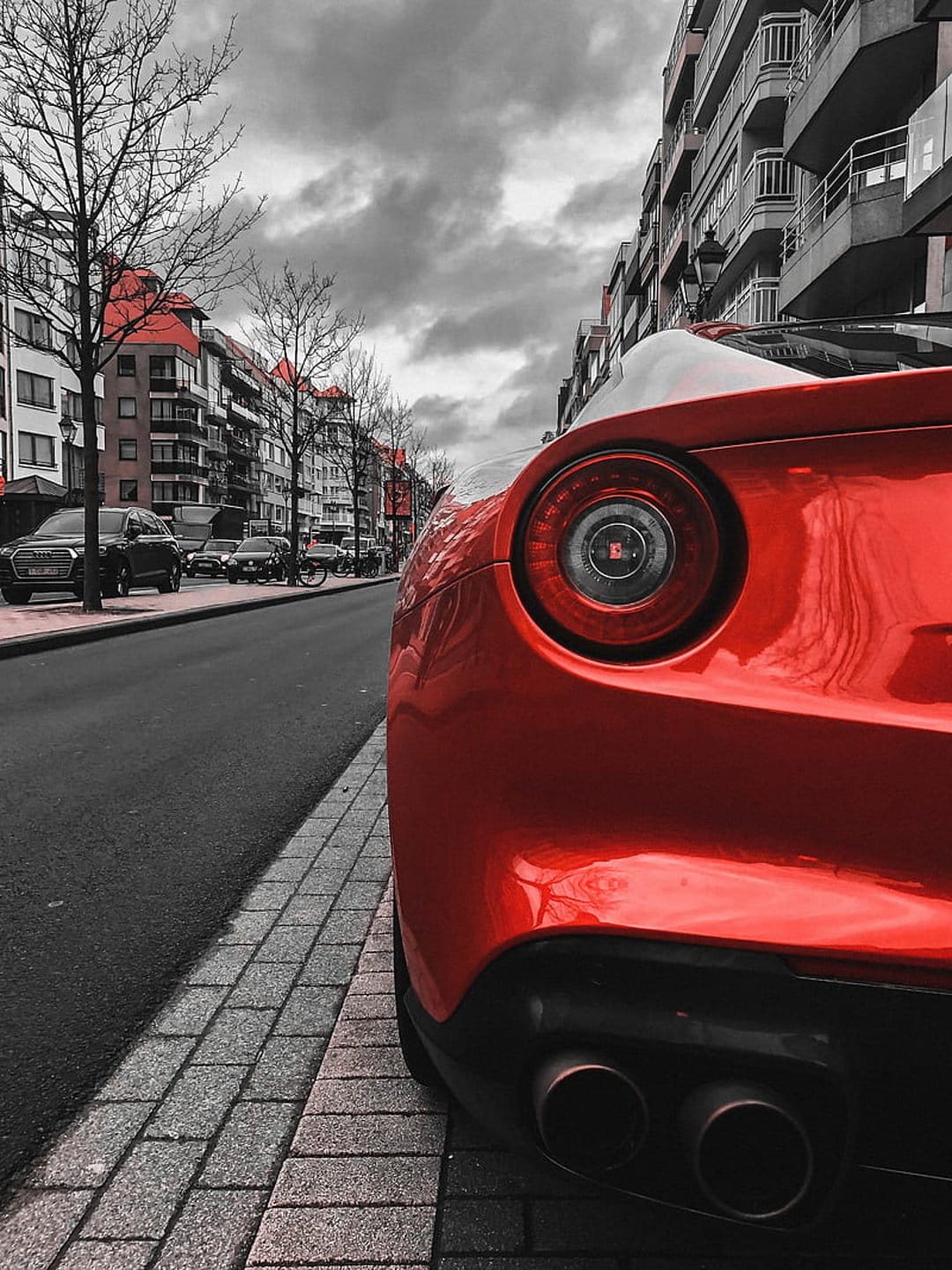 Red Ferrari parked on cobblestones under dark clouds, utilizing selective coloring against a grayscale city.