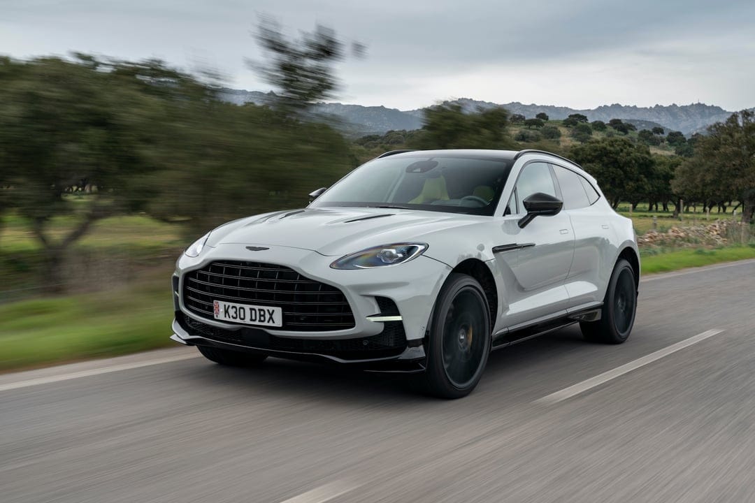 Light grey Aston Martin DBX SUV driving on a paved road with blurred green trees and distant mountains.