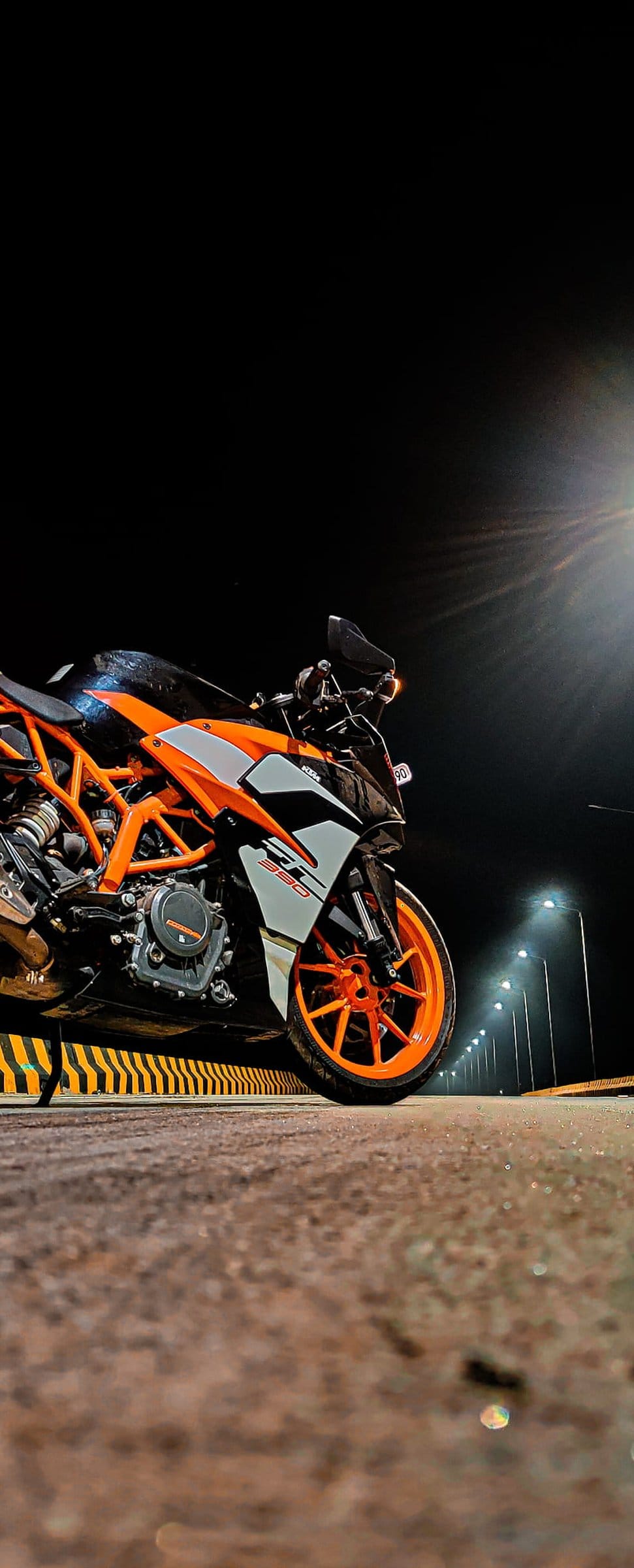 Orange and white KTM motorcycle parked on a dark asphalt road at night with streetlights and a lens flare.