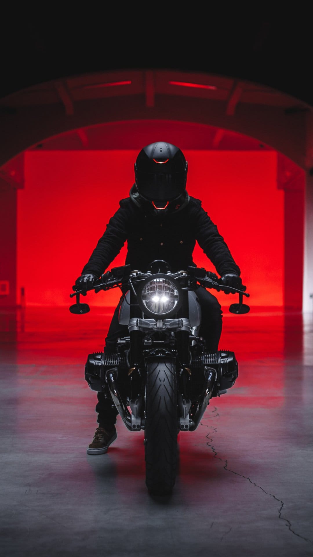 Biker in black gear sits on a motorcycle with a round headlight under intense red lighting on a concrete floor.