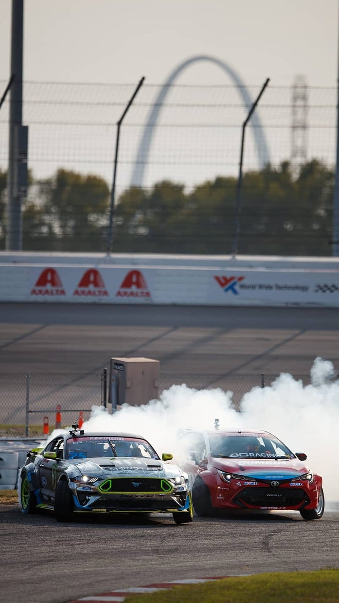 Mustang and Corolla drift cars sliding on a track with thick white smoke against the St. Louis Gateway Arch.