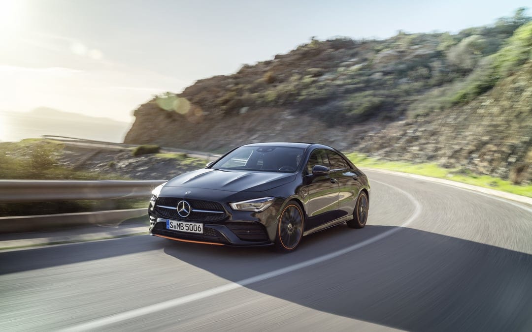 Black Mercedes-Benz CLA coupe with orange accents driving on a winding coastal road with mountain motion blur.