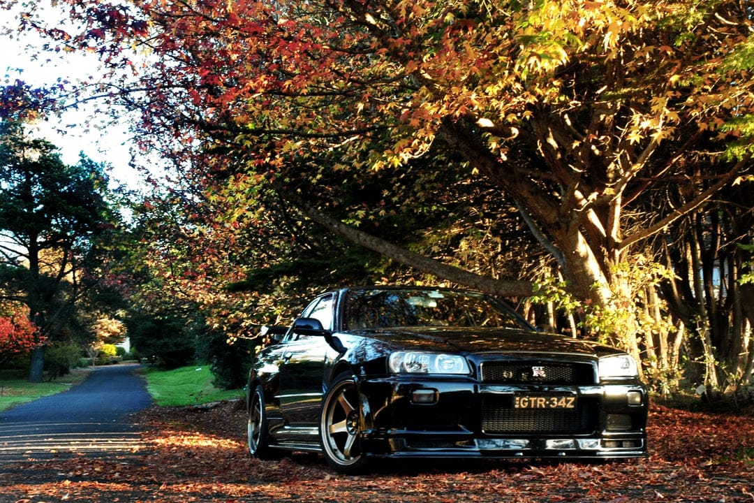Black Nissan Skyline GT-R R34 parked on a road covered in red and orange autumn leaves under large trees.