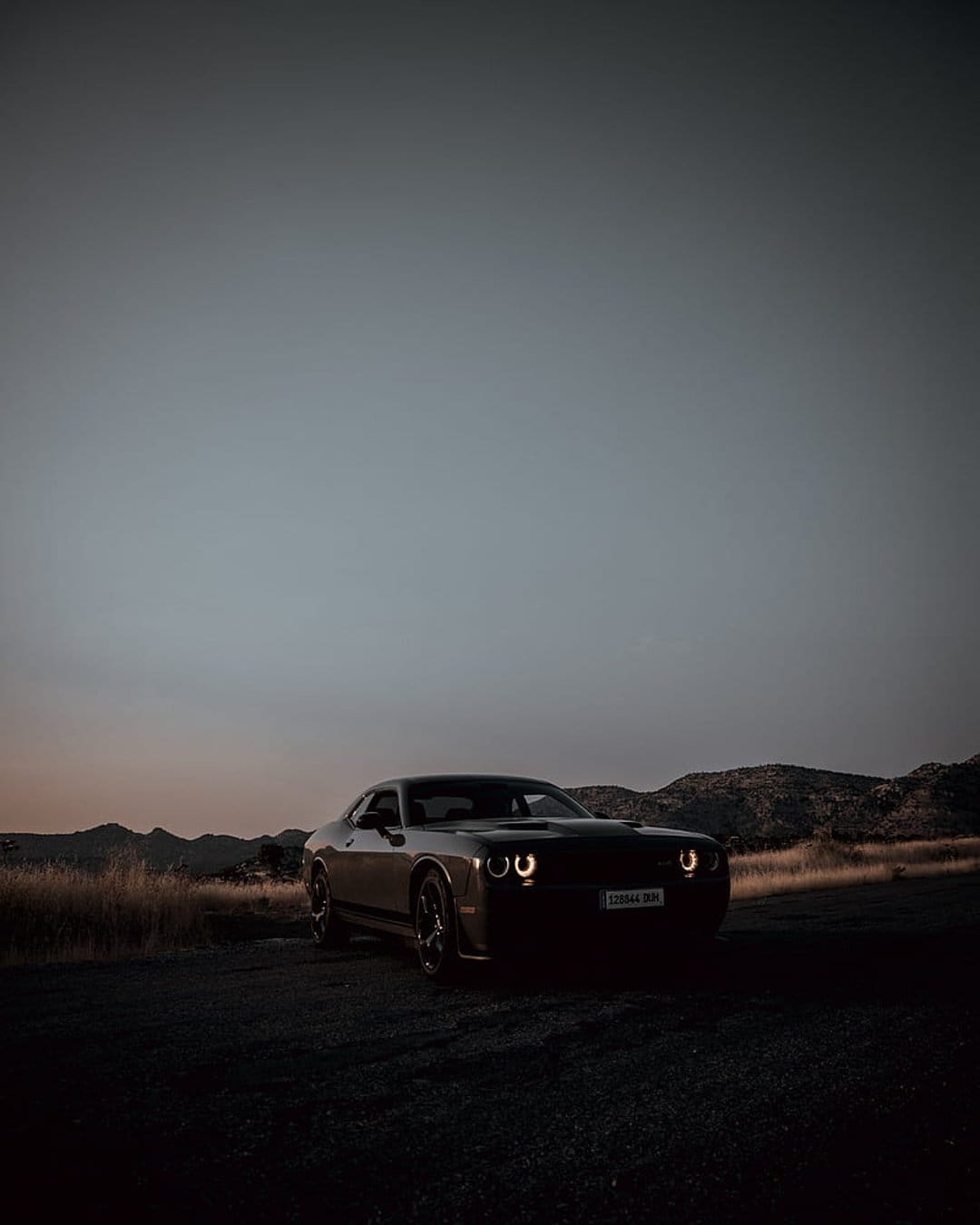 Dodge Challenger with glowing headlights parked on a dirt trail against a sunset mountain range backdrop.