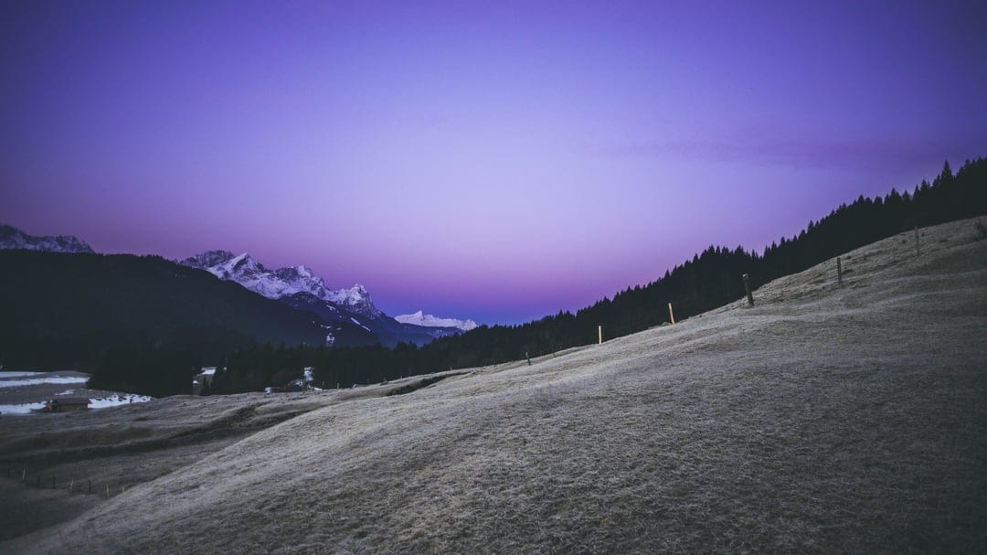 Snow-capped mountains and a grassy hill sit beneath a deep purple twilight sky during a calm alpine evening.