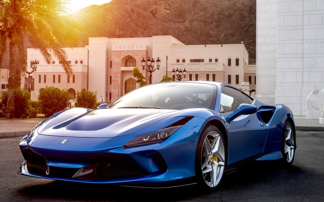 Blue Ferrari sports car parked near an elegant building and mountain during a golden sunset with lens flare.