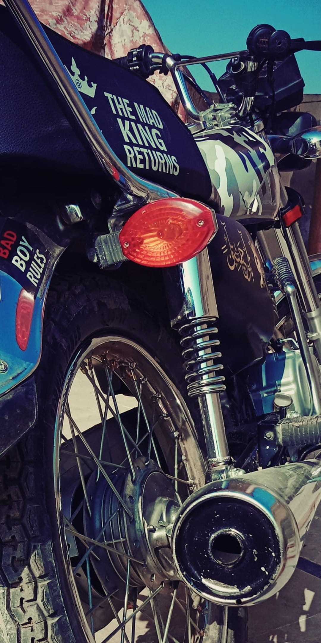 Custom motorcycle rear wheel with a red tail light, chrome shock absorber, and stickers on a blue fender.
