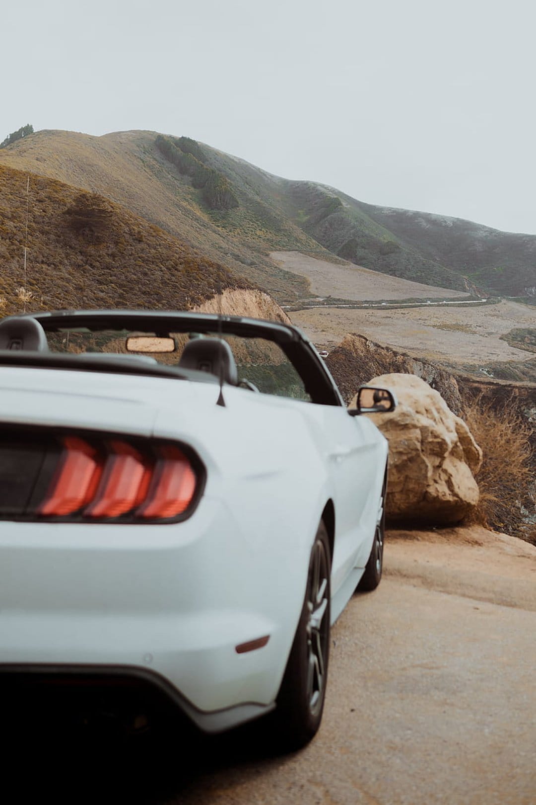 White Ford Mustang convertible parked on a winding mountain ridge featuring iconic red taillights and green valleys.