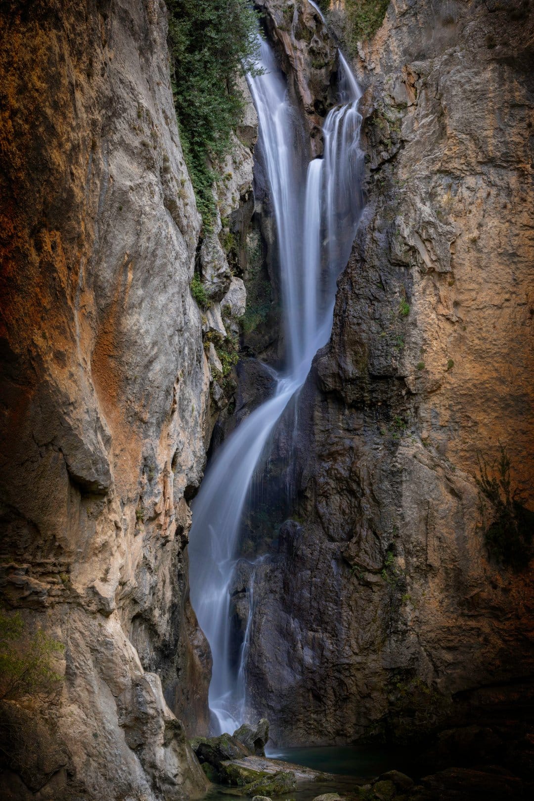 Waterfall with silky white water flows between tall, textured brown canyon walls and green mossy rock ledges.