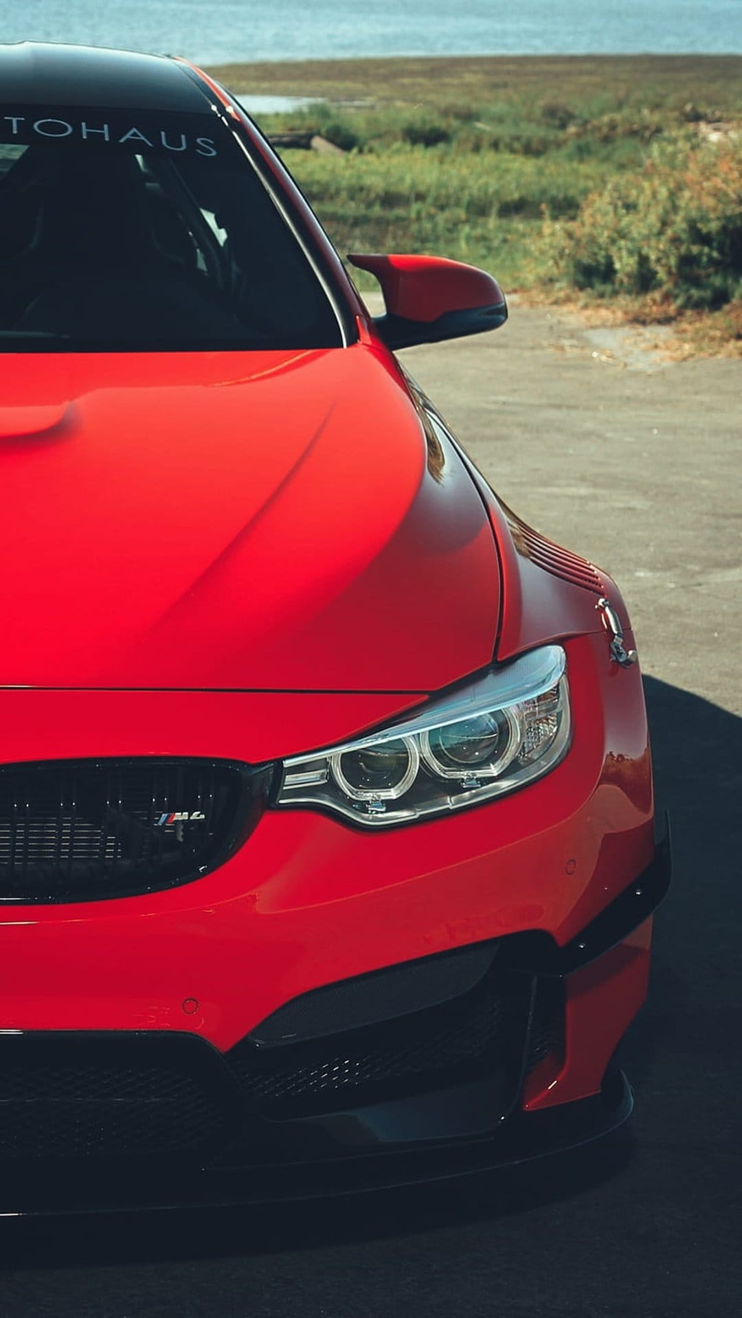 Crimson BMW M4 sports car with bright headlights parked near a tranquil lakefront under clear, sunny summer skies.