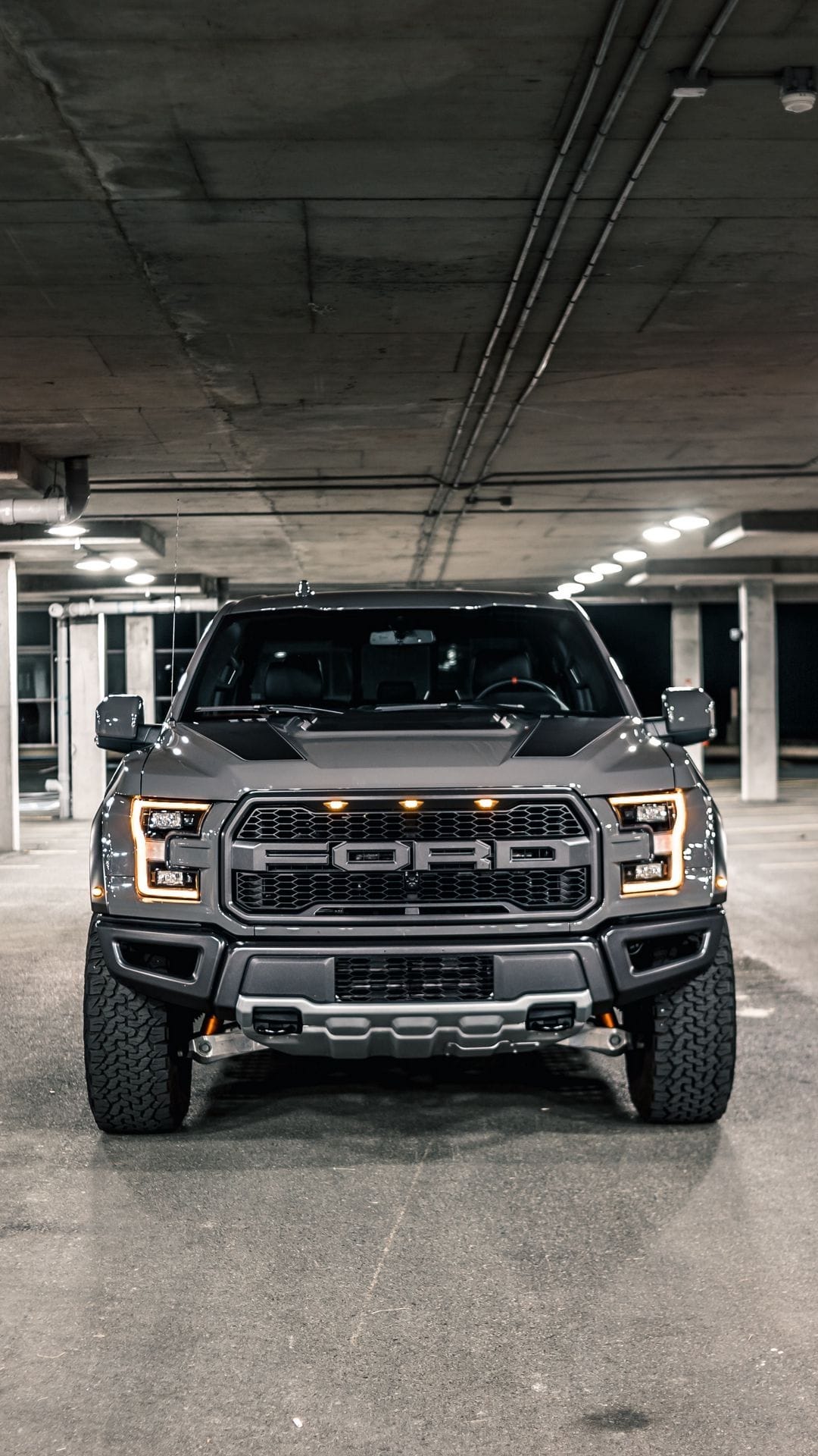 Front view of a grey Ford F-150 Raptor with glowing amber lights in a dark, concrete underground parking garage.