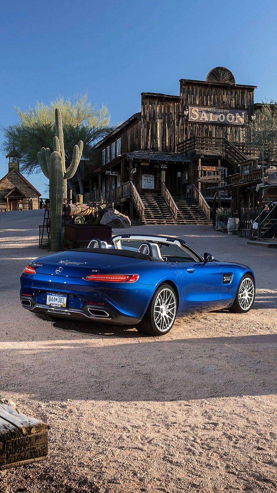 Blue Mercedes convertible parked on a dirt road between rustic wooden saloons and desert cacti under a clear sky.