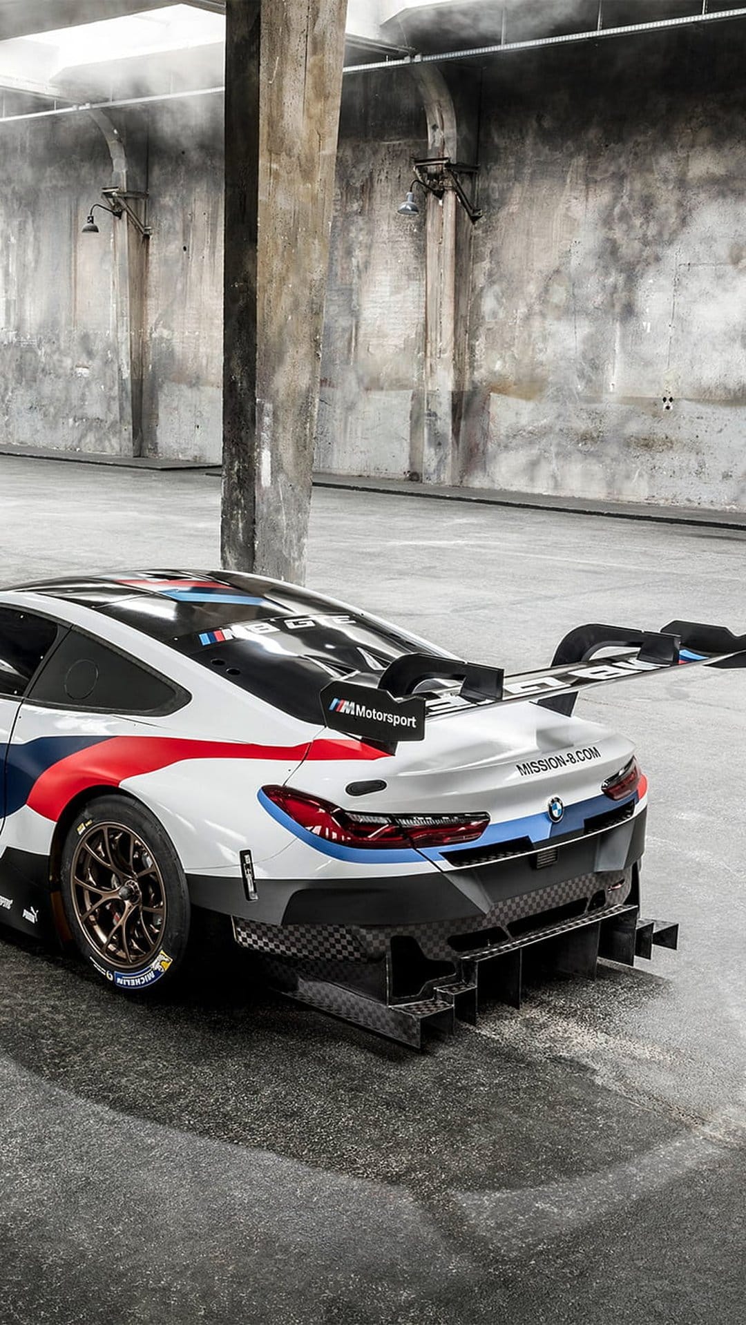 BMW M Motorsport race car with a massive rear wing and red livery parked by concrete pillars in a dark warehouse.