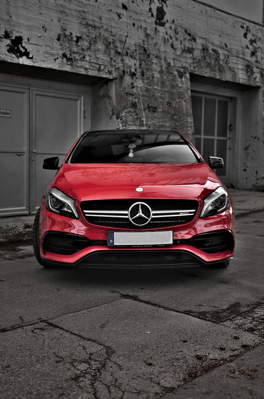 Red Mercedes-AMG A-Class car parked on cracked asphalt in front of a weathered gray industrial building door.