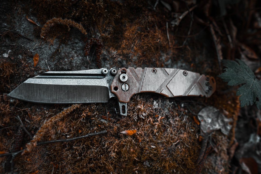 Tactical folding knife with a distressed metal blade resting on a thick bed of green forest moss and damp earth.