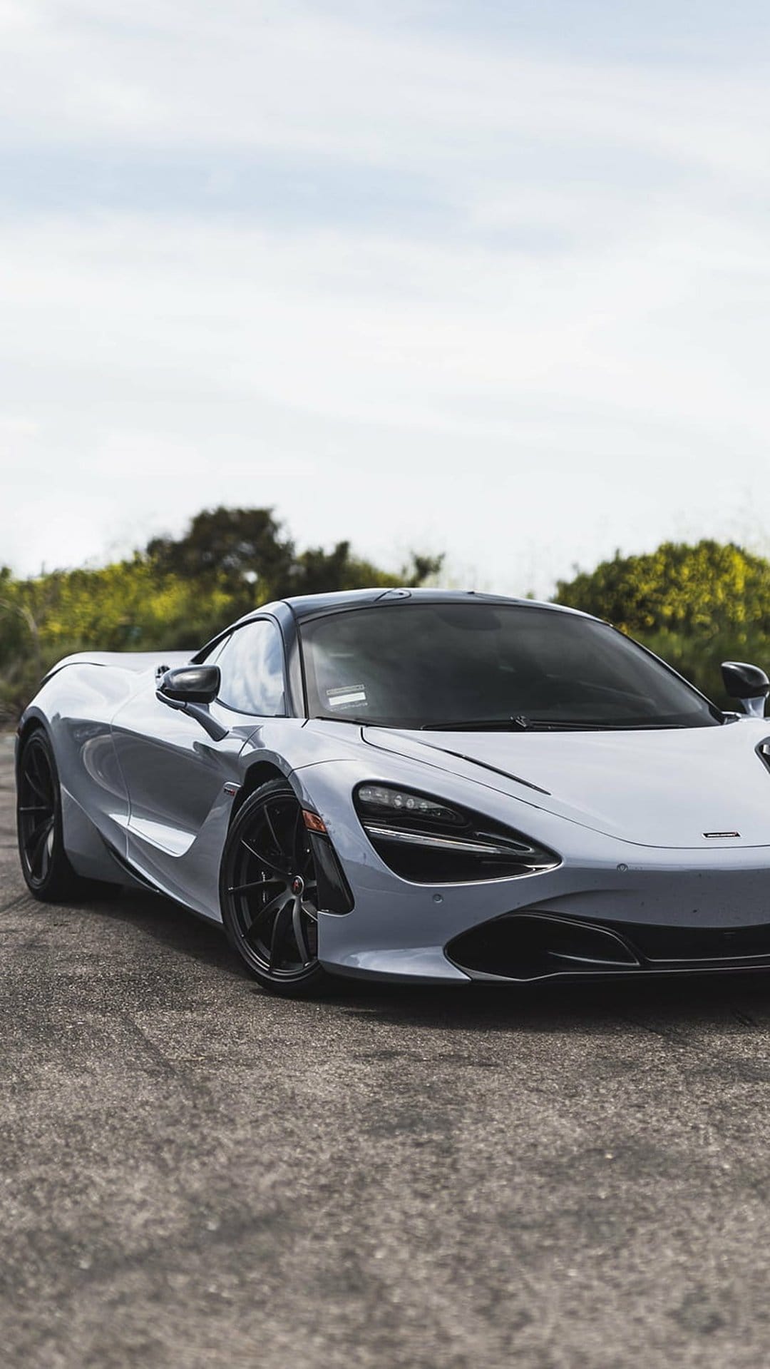 McLaren 720S supercar in metallic grey with black rims parked on asphalt near green trees under a cloudy sky.