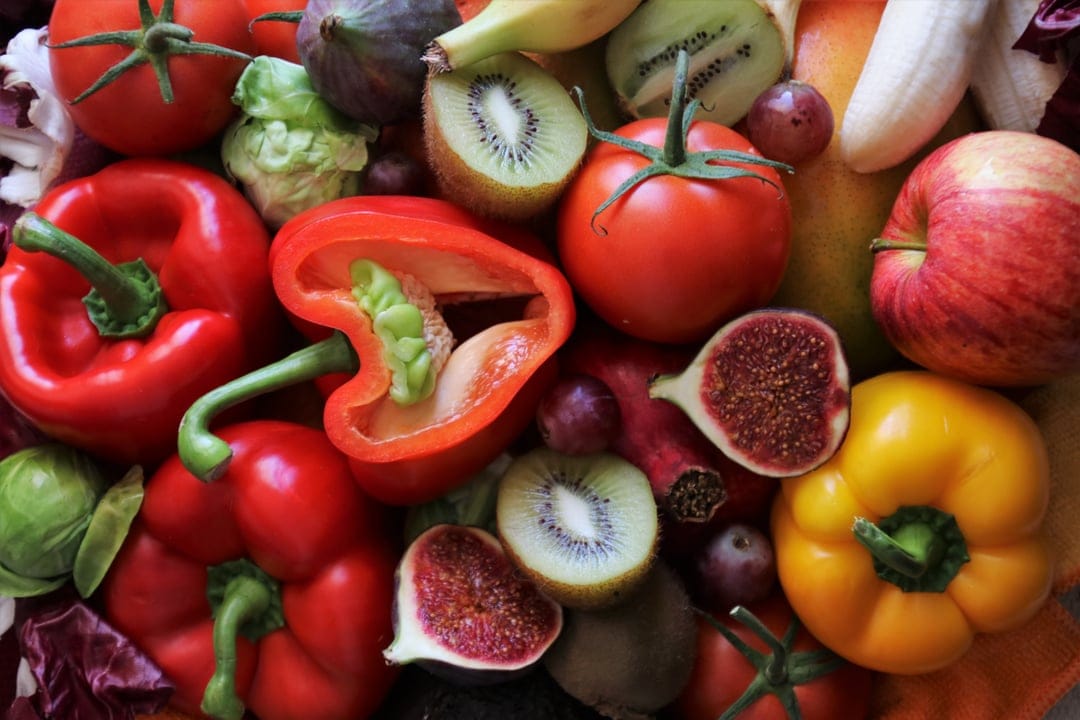 Fresh red bell peppers, sliced kiwi, and purple figs are displayed in a colorful overhead produce arrangement.