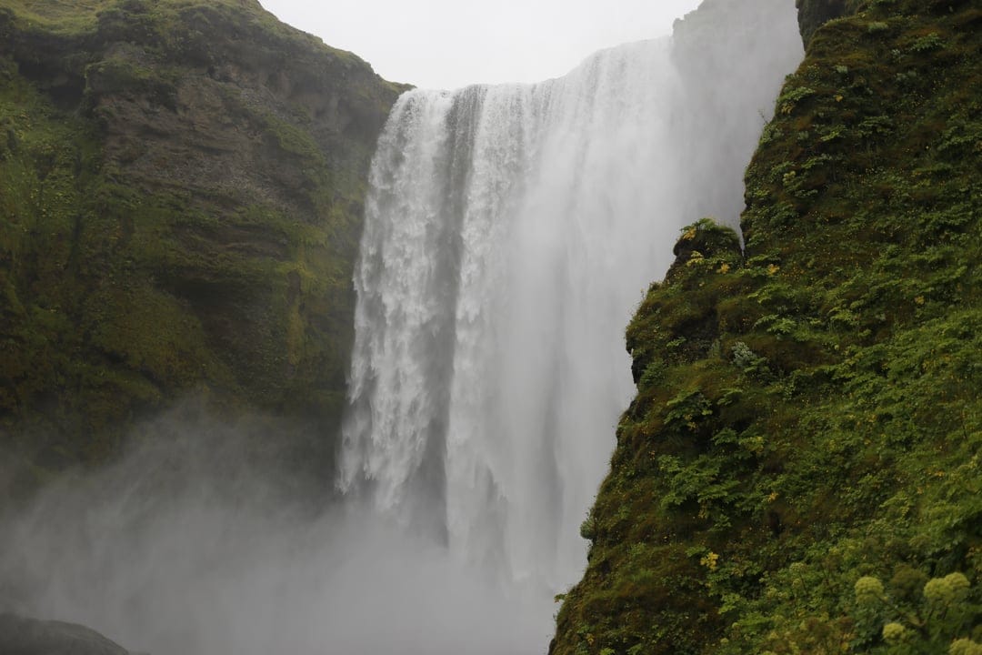 Skogafoss waterfall drops from mossy green cliffs into a misty pool within a vertical Icelandic valley landscape.
