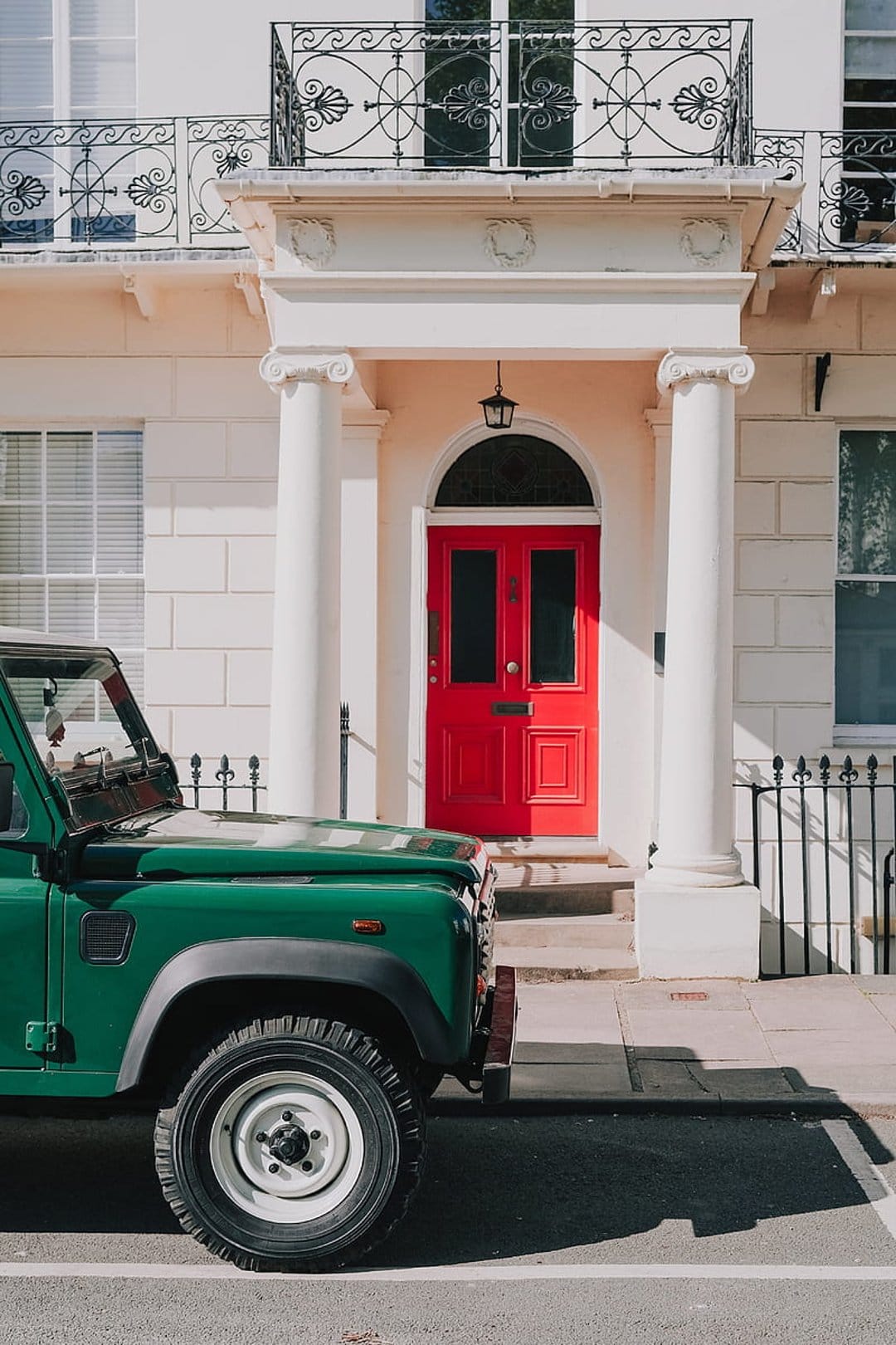 Forest green Land Rover Defender parked on a sunny street in front of a white building with a bright red door.