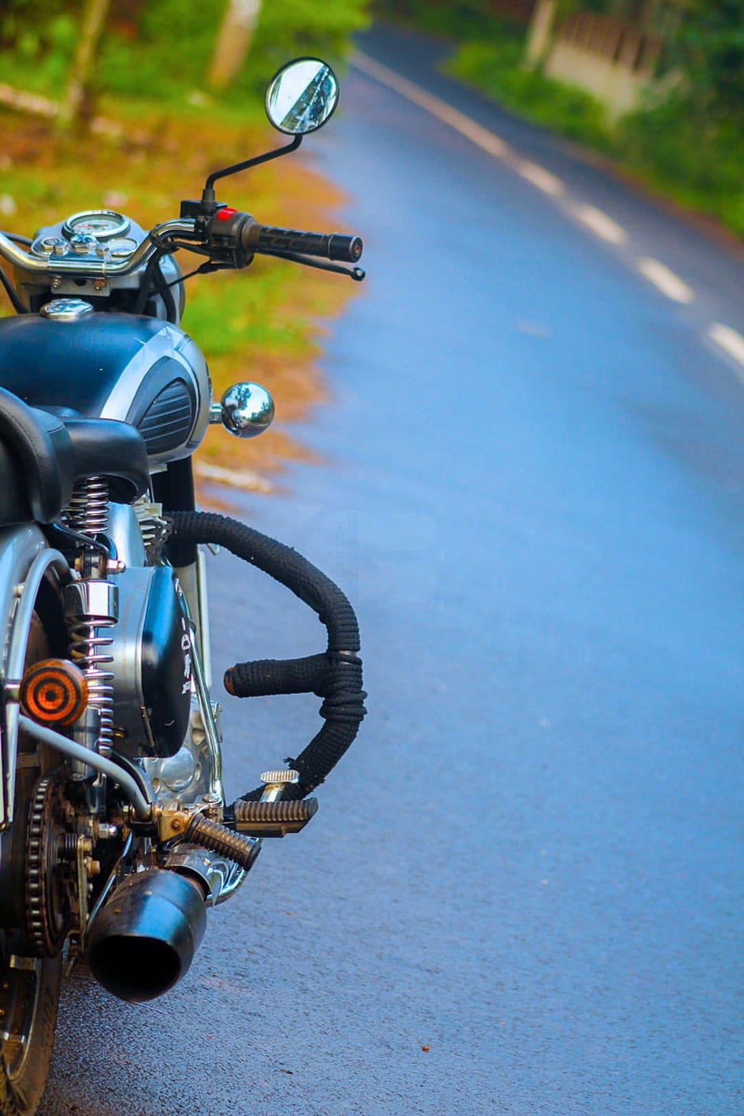 Classic motorcycle handlebars and chrome engine parts focus on a wet asphalt road with a blurred green background.