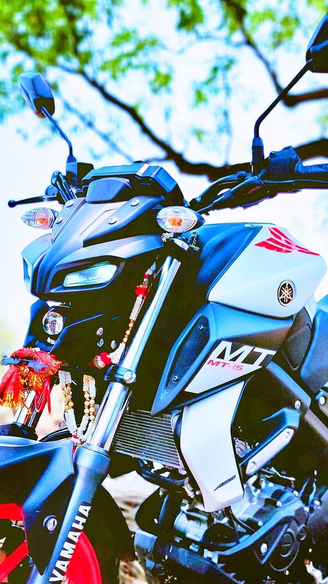 Yamaha MT-15 motorcycle front headlamp and handlebars with decorative strings against a blurred green tree canopy.