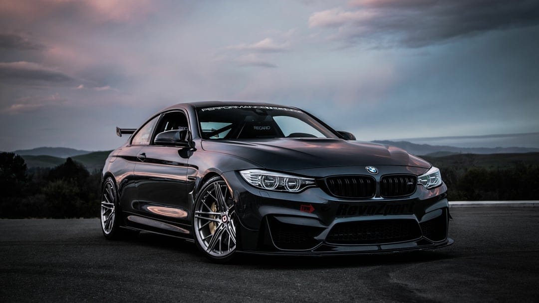 Dark grey BMW M4 with carbon fiber aero and racing wheels parked on asphalt against a sunset sky.