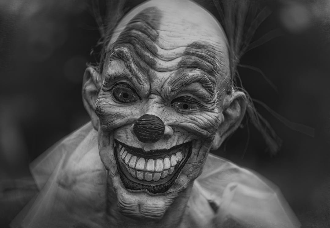 Creepy clown mask with sharp teeth, wrinkled skin, and wild hair in a high-contrast black and white close-up.