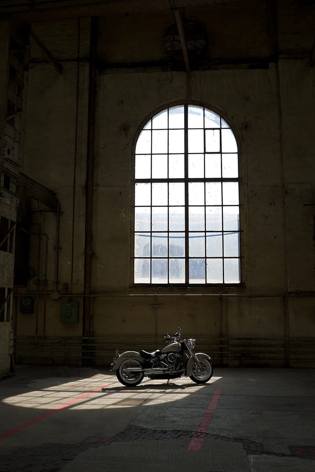 Retro motorcycle parked on a concrete floor inside a dark warehouse with sunlight streaming from an arched window.