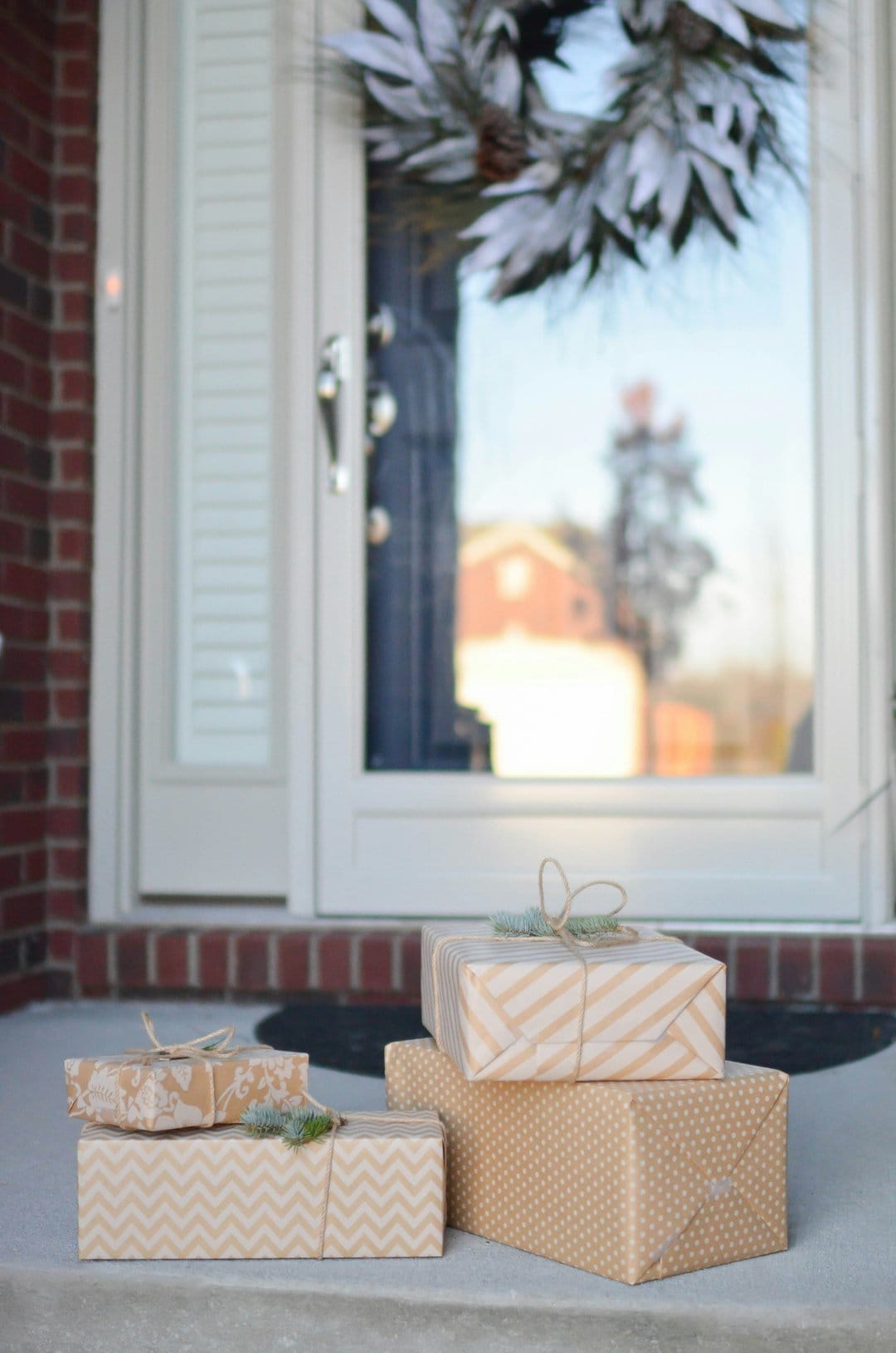 Stacked holiday gifts with twine bows sit on a porch in front of a glass door with a frosted winter wreath.