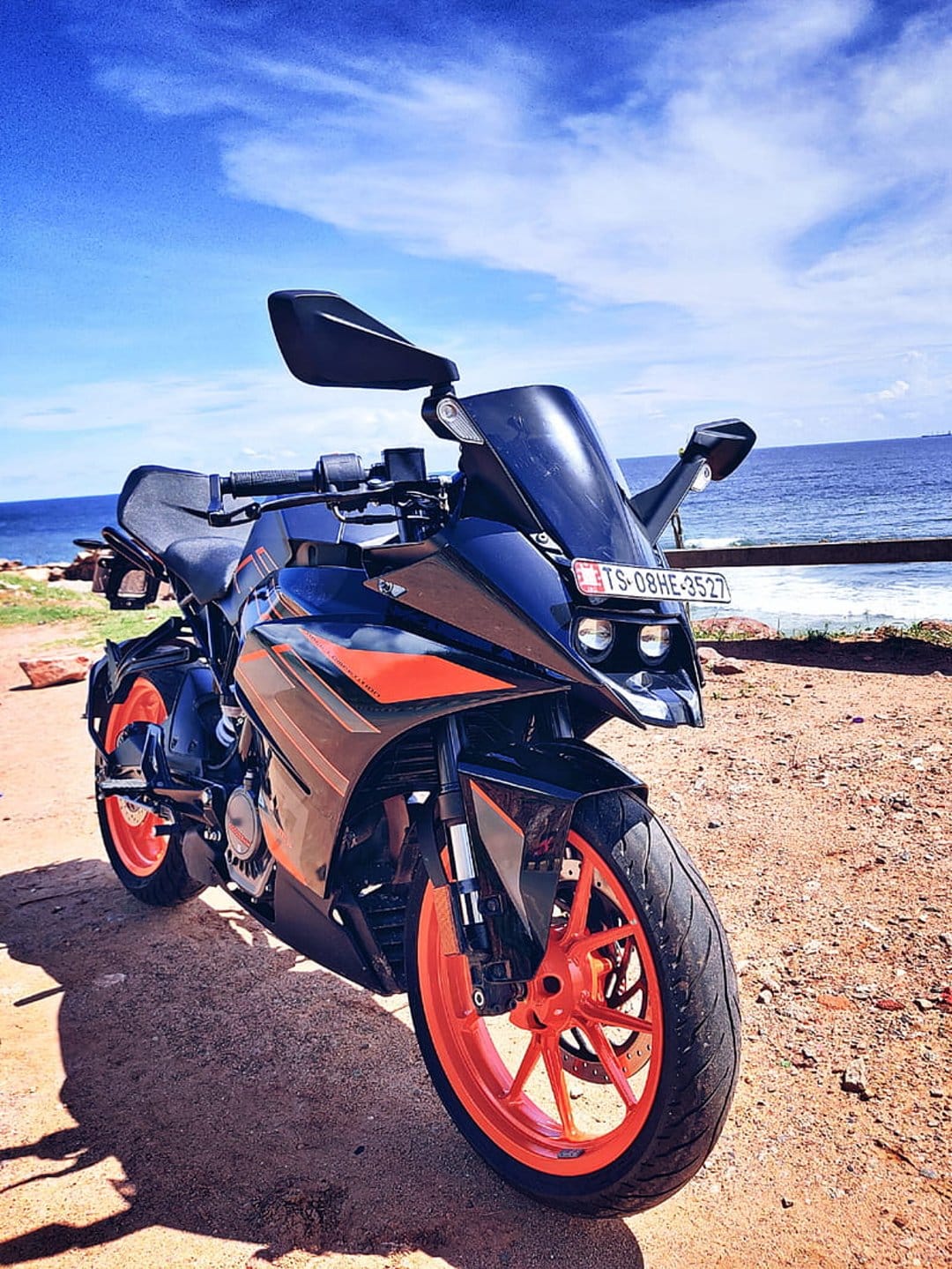 Orange and black KTM motorcycle parked on a sandy beach with blue ocean waves and a clear sky in the background.