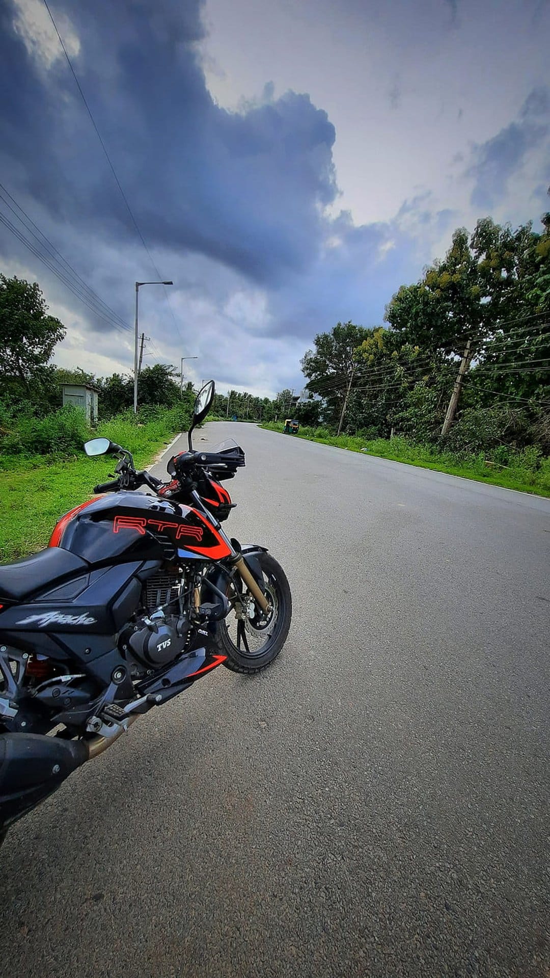 TVS Apache RTR sportbike parked on an asphalt roadside under a dark, dramatic sky with lush green trees behind.