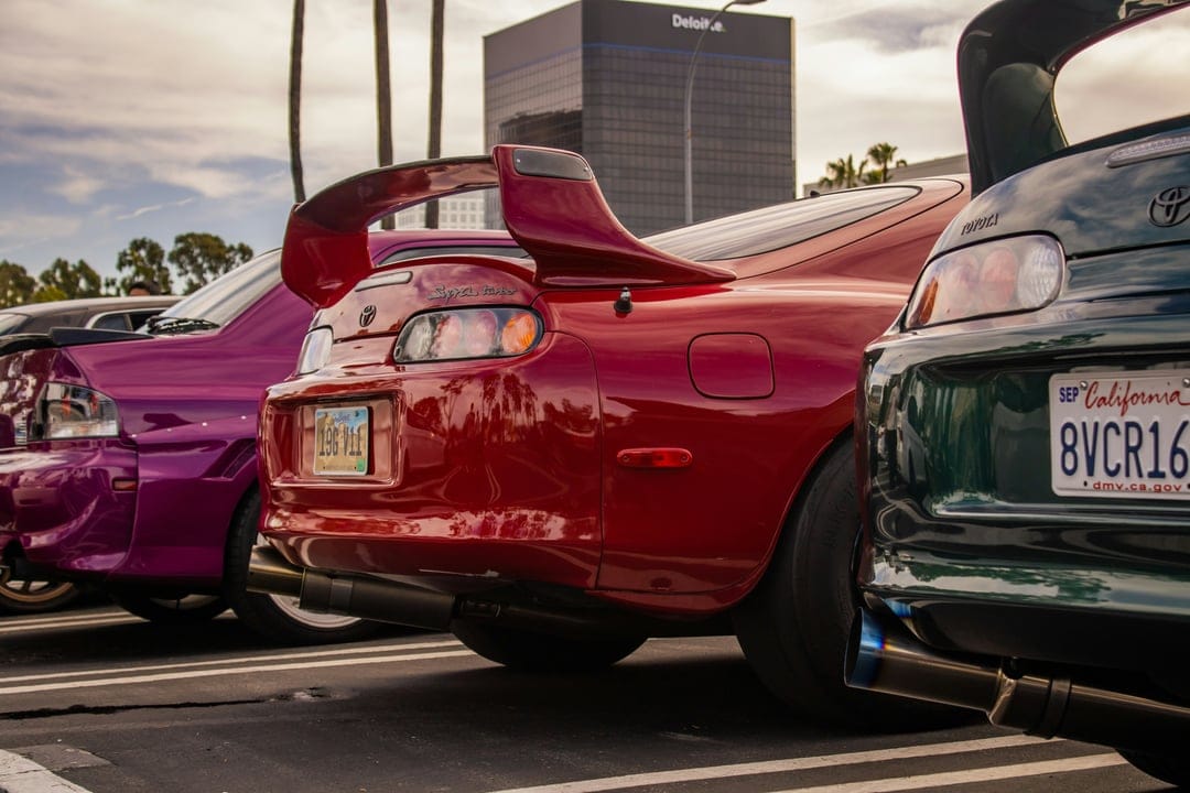 Red Toyota Supra and custom tuner cars parked in a row against a backdrop of palm trees and modern architecture.