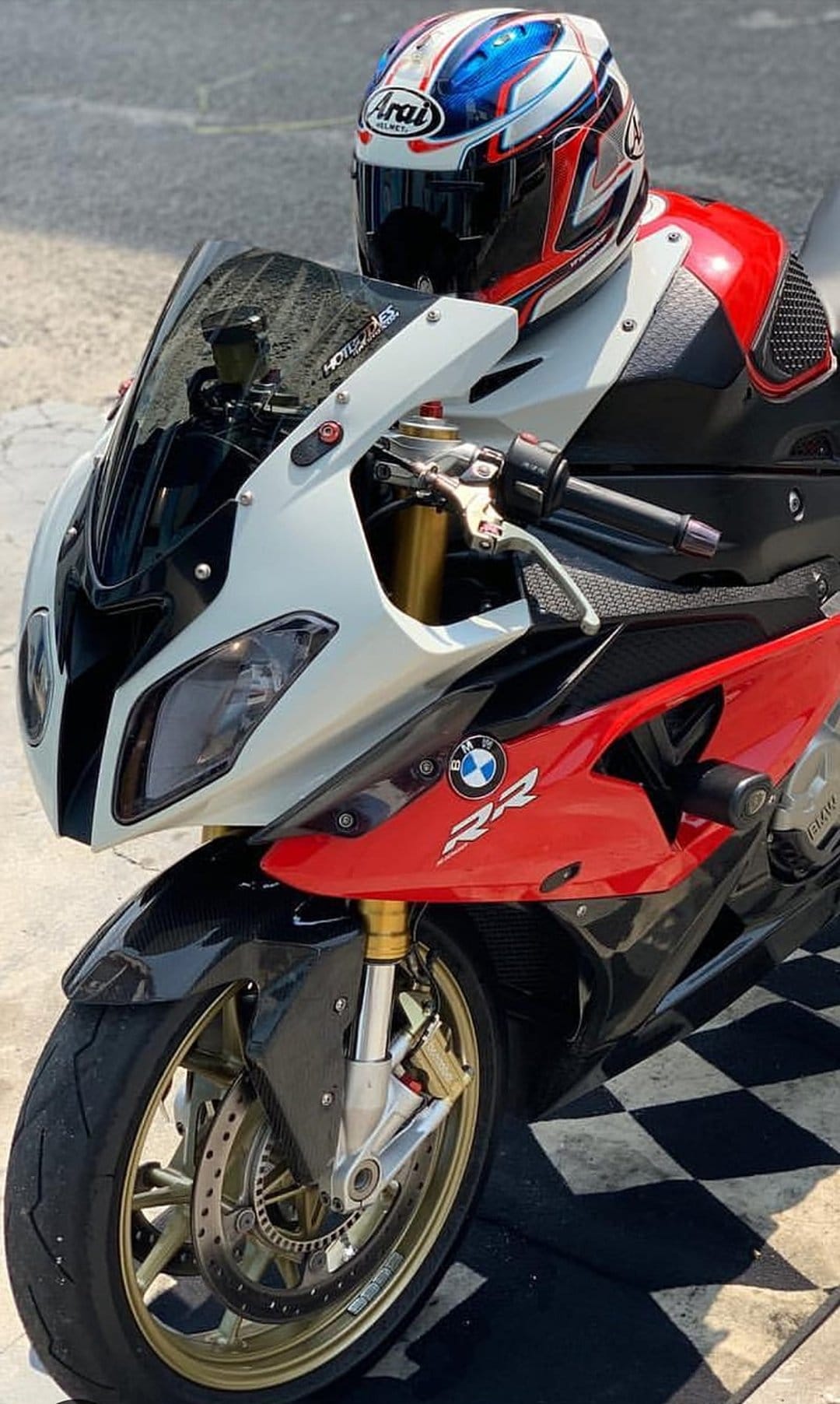 BMW S1000RR motorcycle with red and white fairings rests near an Arai helmet on a black and white checkered floor.