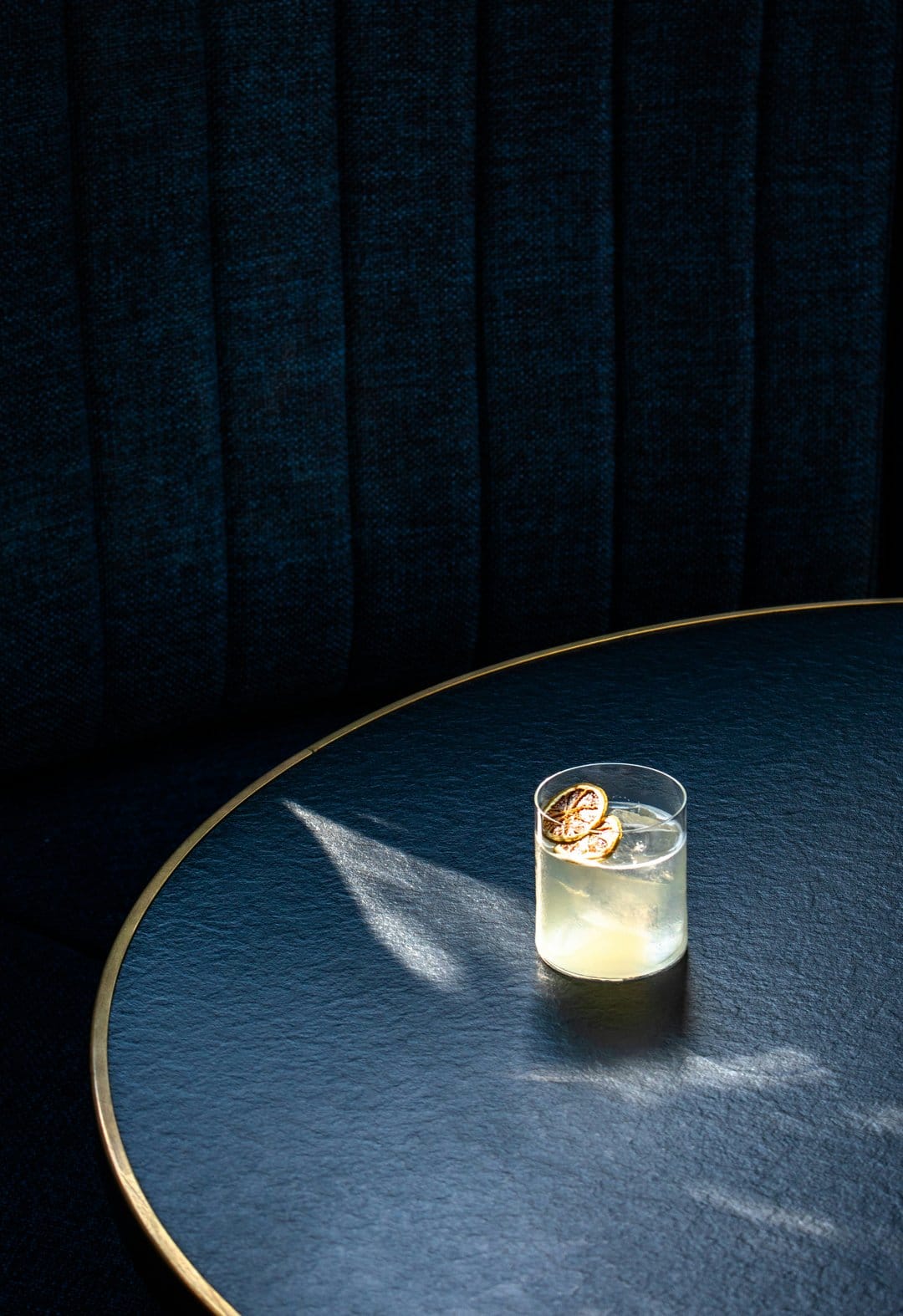 Clear cocktail glass with two dried lemon slices on a dark blue table with gold trim and tufted fabric background.