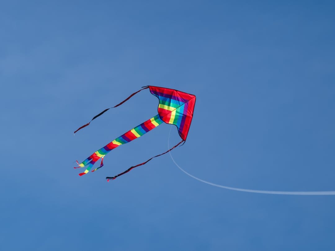 Rainbow-striped delta kite with a long flowing tail soaring across a vast and empty bright blue summer sky.