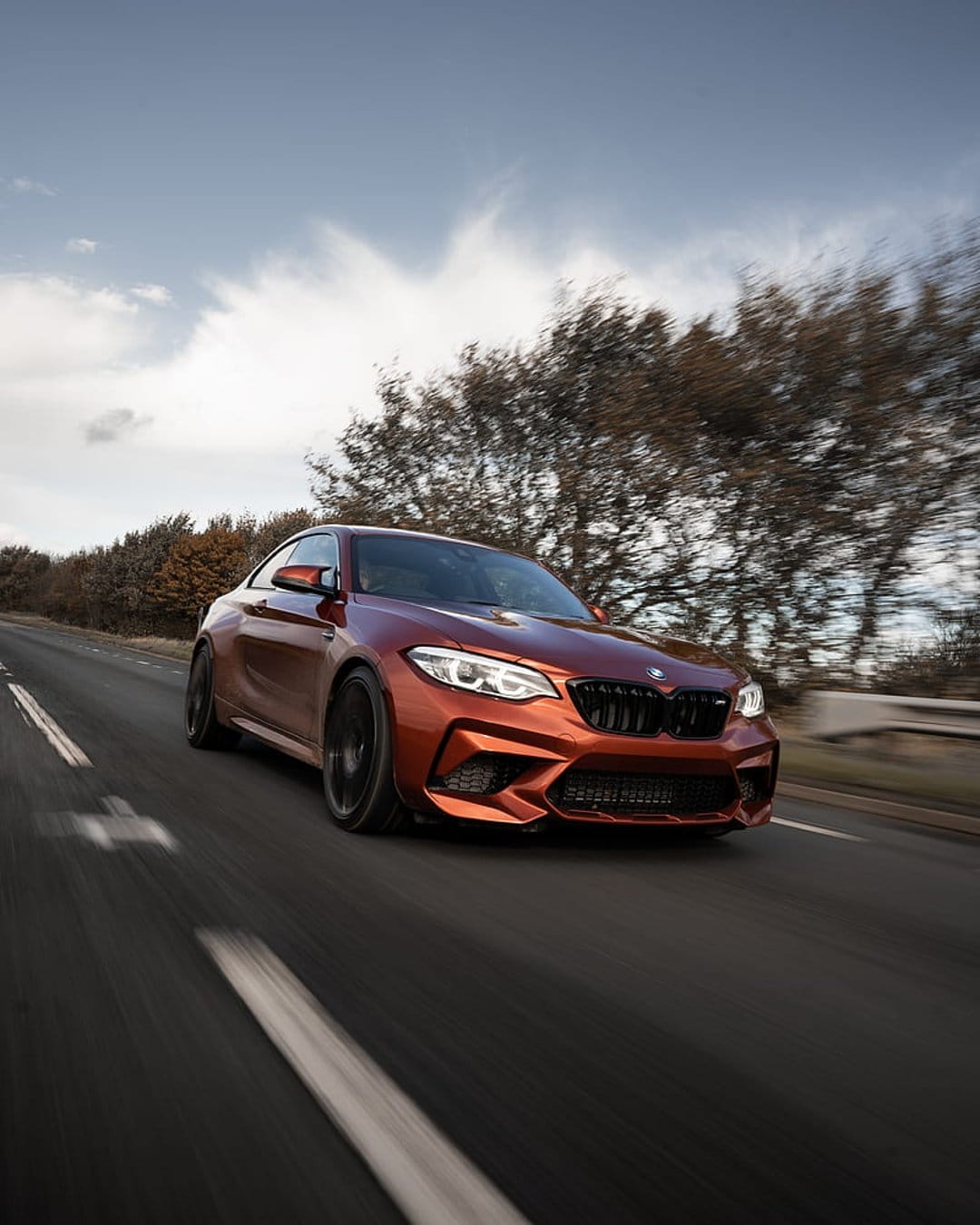 Orange BMW M2 coupe speeds along a highway with motion blur against a background of trees and a cloudy sky.