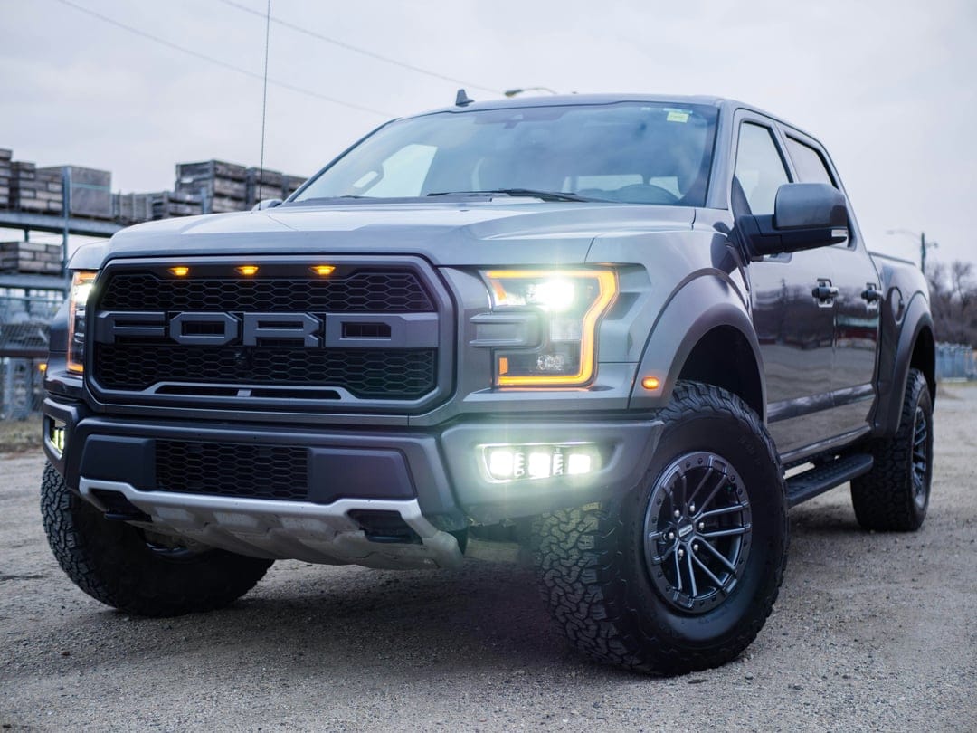 Gray Ford Raptor pickup truck front view showcasing illuminated LED headlights and a bold black mesh grille.