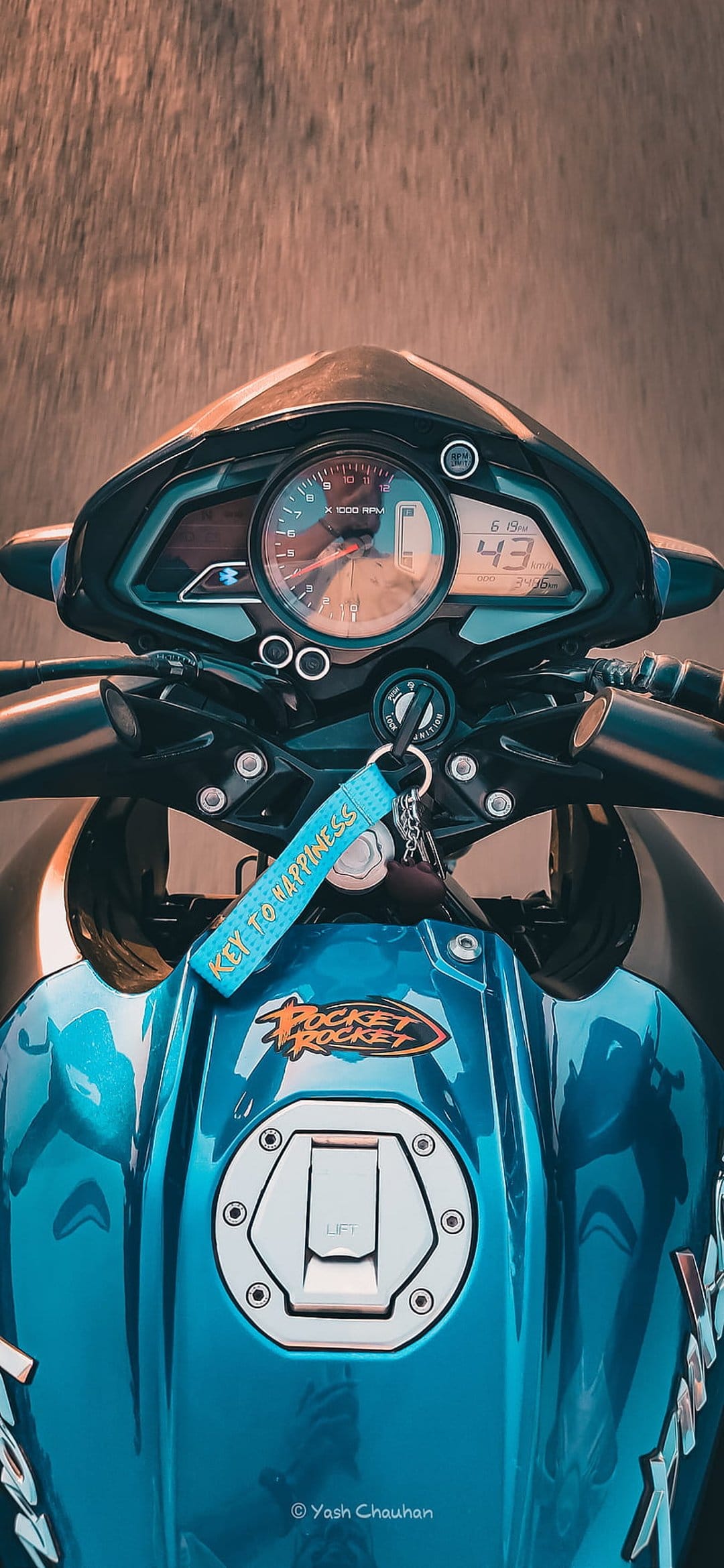 Blue motorcycle dashboard with a Key to Happiness keychain hanging near the speedometer and fuel tank emblem.