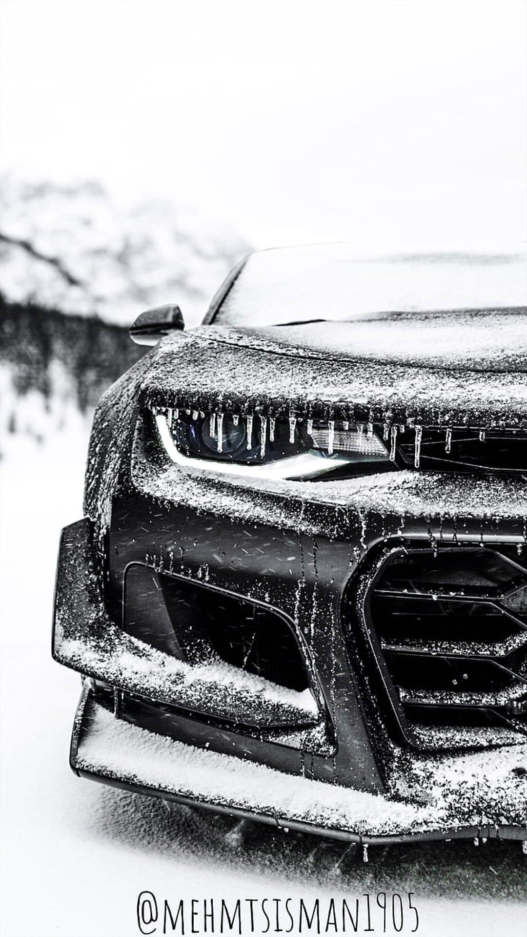 Black car front grille and headlights covered in sharp icicles and thick snow against a blurred winter scene.