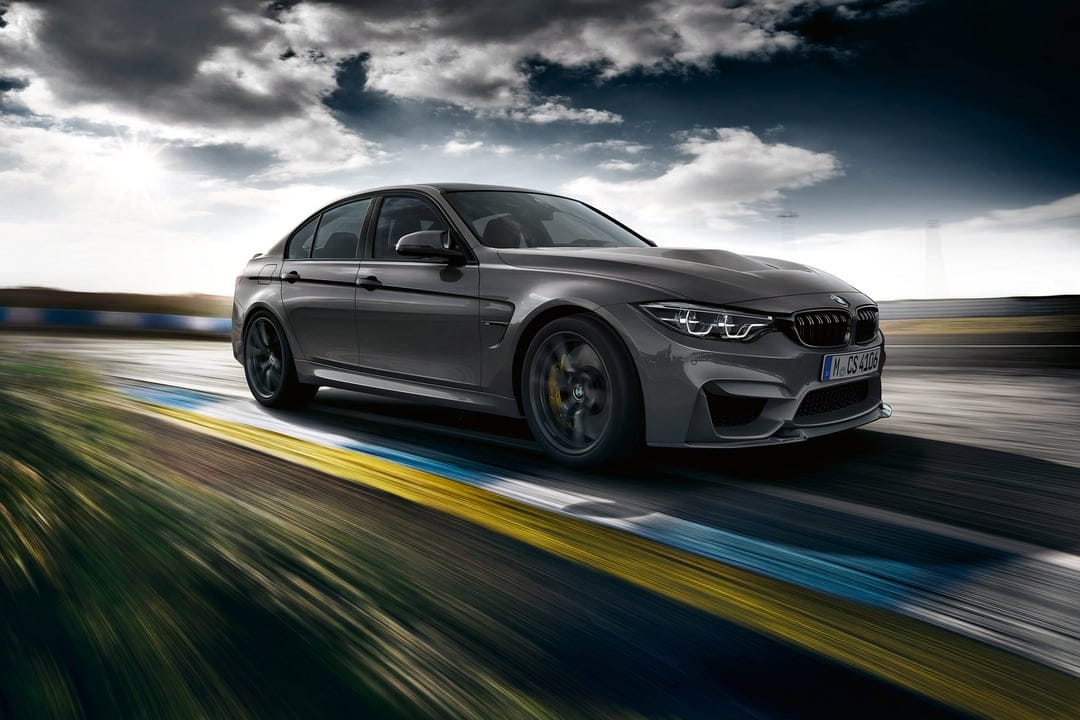 Grey BMW M3 CS performance sedan driving fast on a racetrack with motion blur under a dramatic cloudy sky.