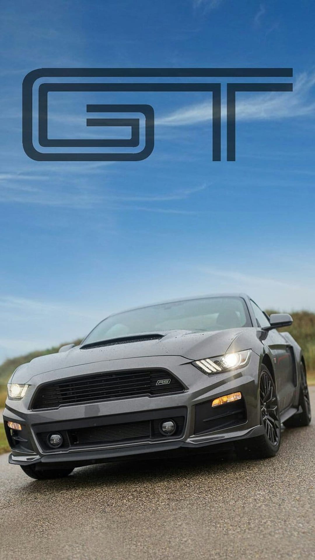 Gray Ford Mustang GT on a wet road with glowing headlights under a blue sky with a GT logo at the top.