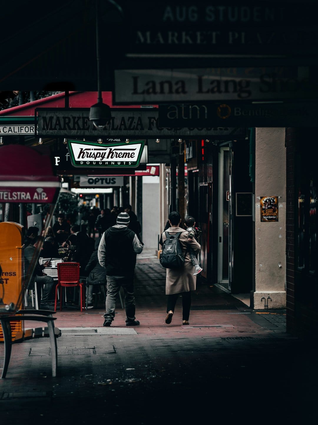 A person carrying a child walks through a dark city alley illuminated by a glowing Krispy Kreme neon sign.