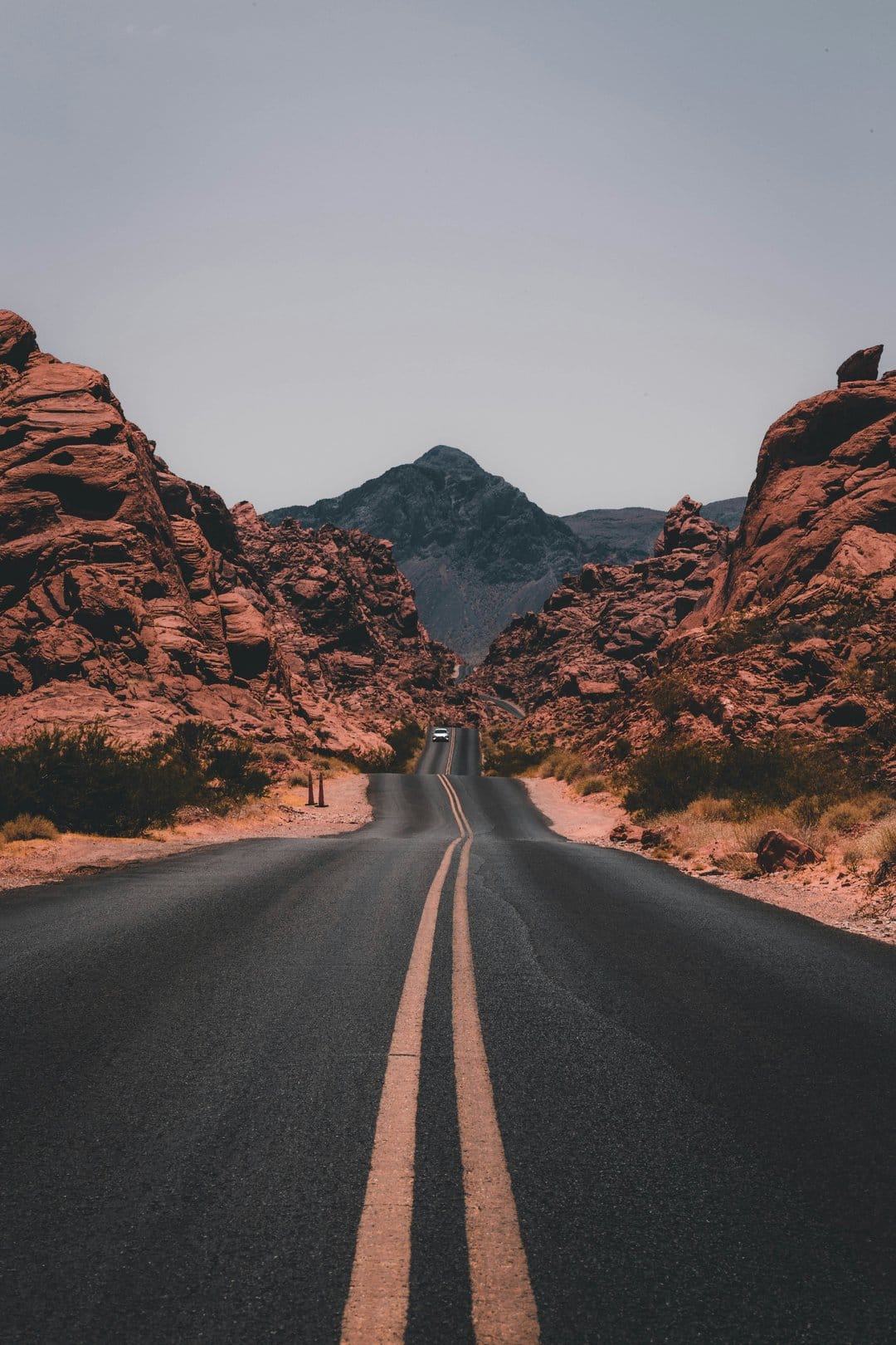 Winding asphalt road with double yellow lines leads past towering red rock formations toward a distant mountain.