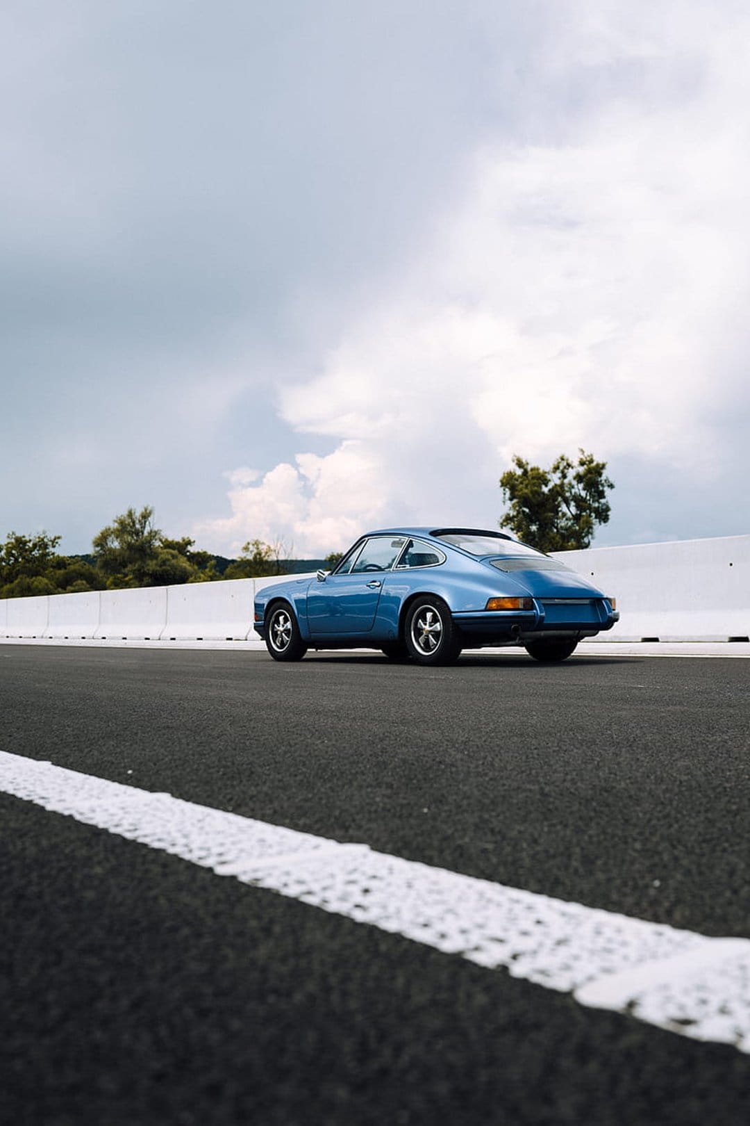 Blue vintage Porsche 911 drives on a wet asphalt road with white markings under a cloudy sky and green trees.