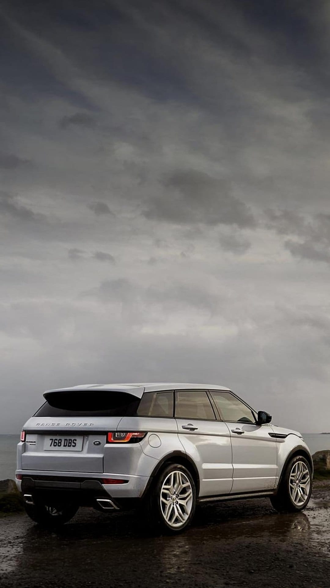 Silver Range Rover Evoque SUV parked on wet pavement beneath a dark, overcast sky showing rear taillights.