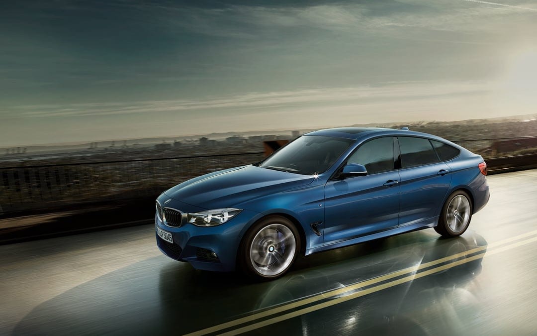 Blue BMW 3 Series GT driving on a wet road at sunset with motion blurred wheels and a distant city skyline.